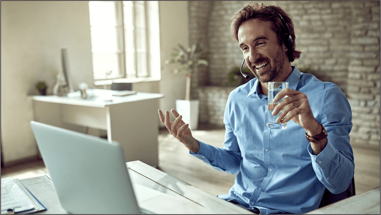 Homem sorridente com fones de ouvido, segurando um copo de água e utilizando notebook